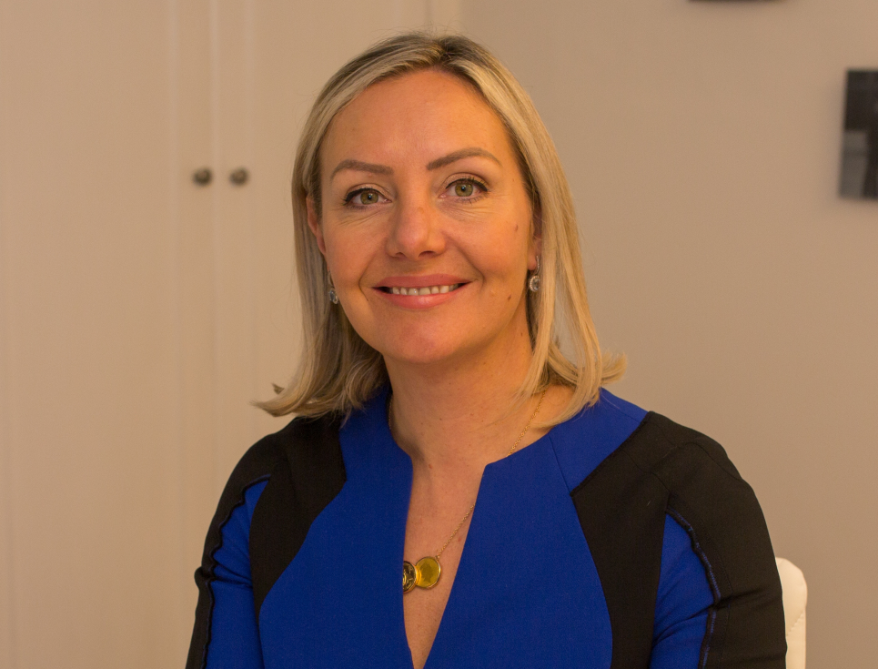 A person with short, blonde hair smiles. They wear a blue and black top.