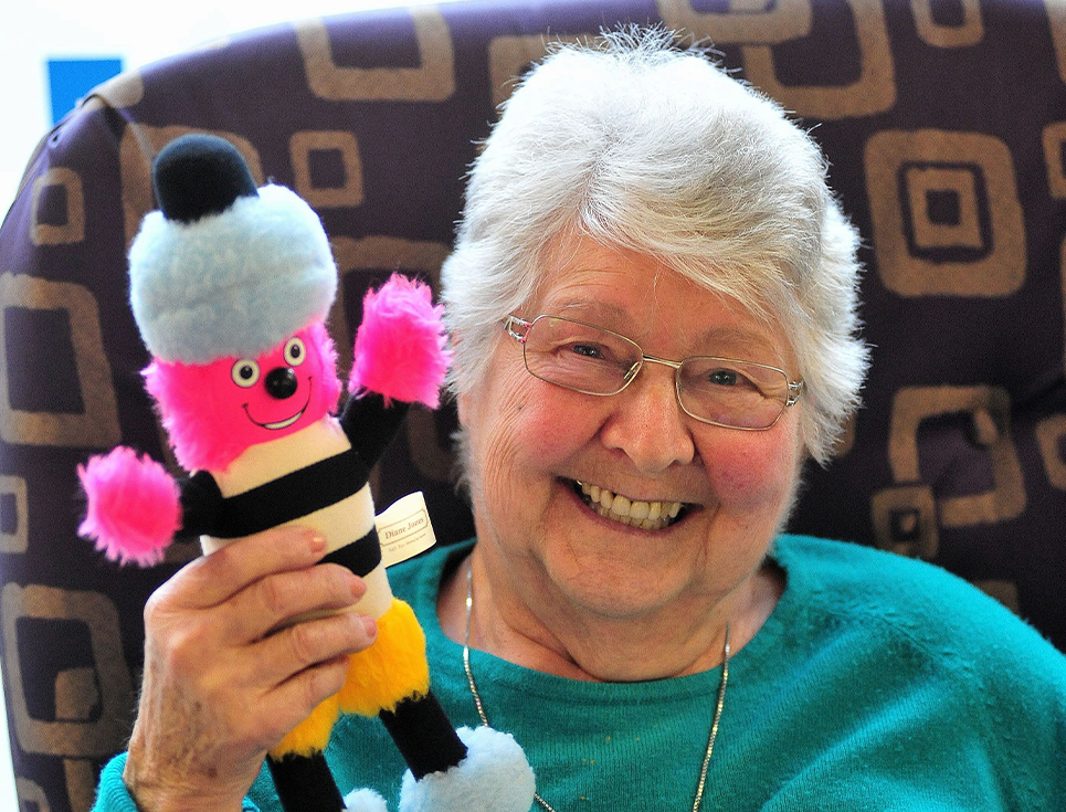 A woman wearing a green jumper holds a soft toy of Bertie Bassett
