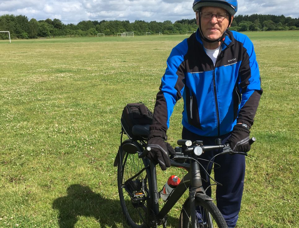 A man stands in a green, grassy field with a black bicycle. He is wearing a blue cycling jacket, black trousers and a blue cycle helmet.