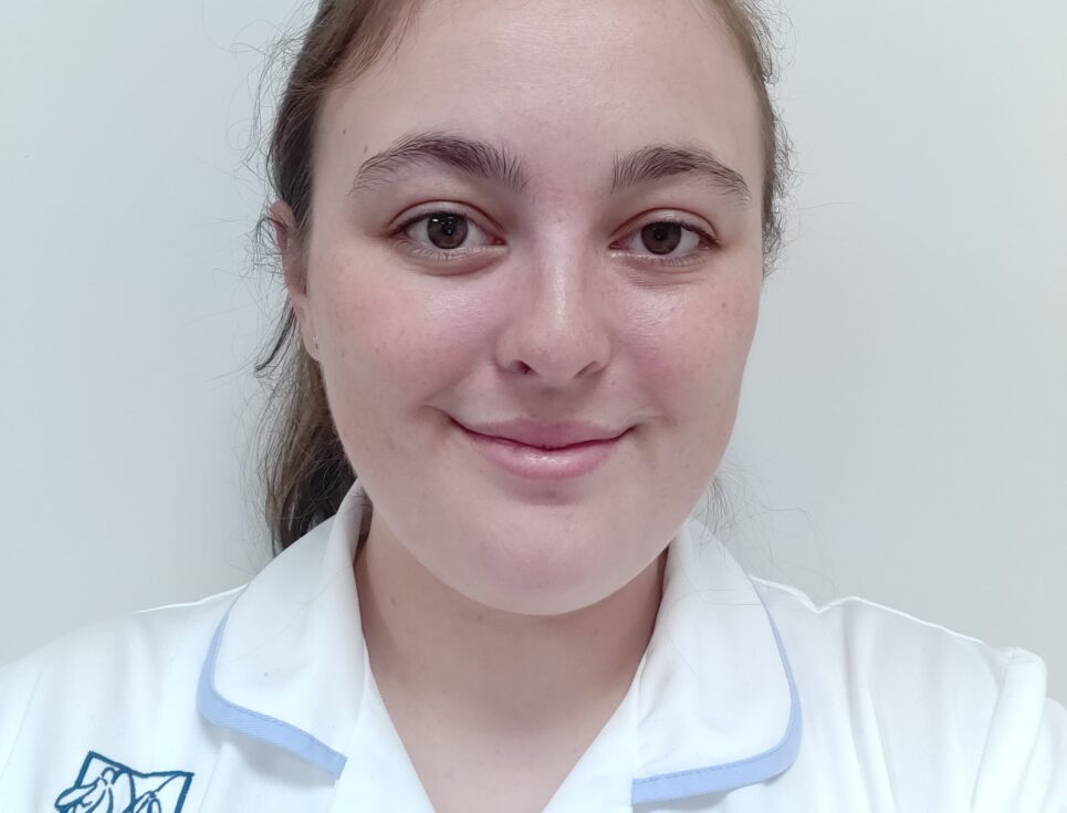 Tia Mountain pictured wearing a white therapy support assistant uniform. She is pictured against a white background and smiling.