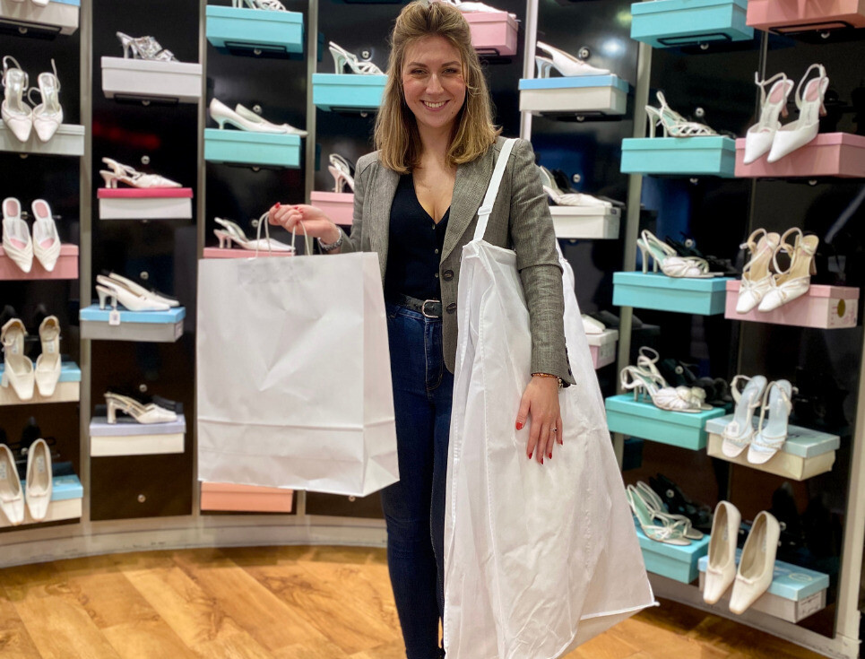 A bride to be holds up a dress bag against a backdrop of a wall of shoes