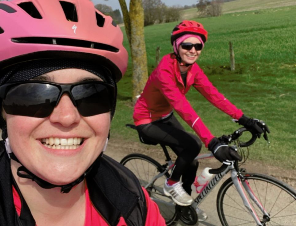 Hannah and Natalie cycle along a country road wearing pink and black riding gear and helmets. They are both smiling.