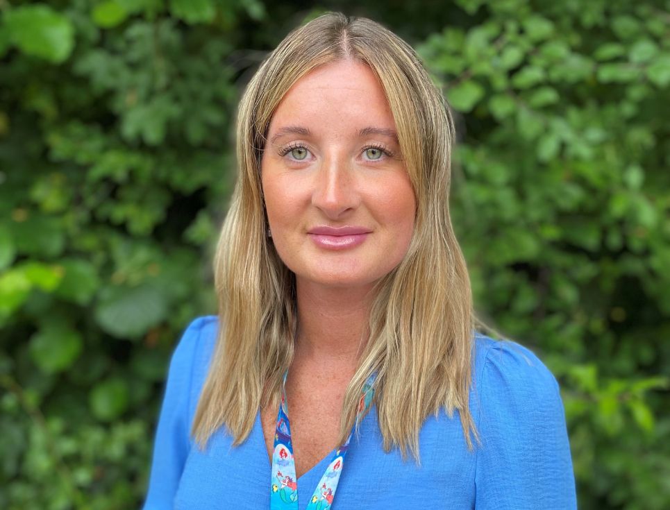 Nikki pictured in front of a dark green leafy bush. She has shoulder length blonde hair and is wearing a bright blue top.