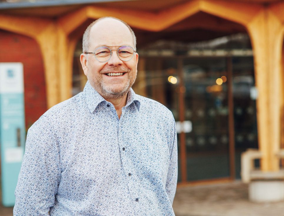 Mike Wilkerson stands outside the front entrance to St Richard's Hospice. He is wearing a pale blue shirt and no tie. He is smiling. Behind him is the welcoming, wood-beamed walkway into the hospice.