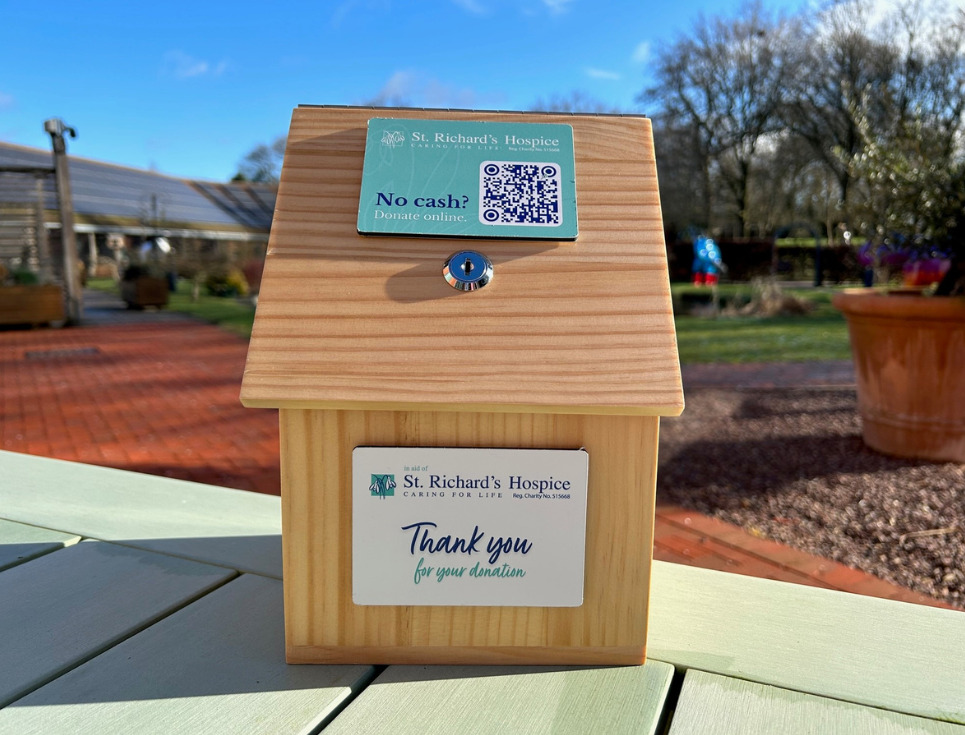 A wooden donation box sits on a wooden table in the hospice gardens. A message on the box reads thank you for your donation.