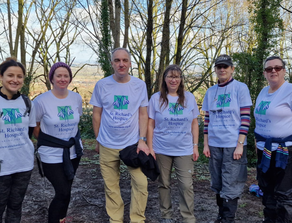 A group of people stand together during a training walk for the Trek to Petra challenge. They are standing in woodland and everyone is wearing white t-shirts featuring the hospice's logo.