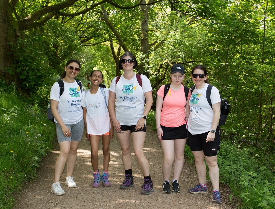 A group of people wearing St Richard's Hospice branded t-shirts stand together during the hospice's Malvern Hills Walk. They are on a pathway through a wooded area with a green canopy of trees.