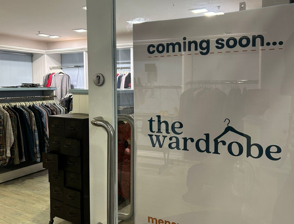 A close-up of an open door leading into the hospice's pop-up menswear shop. A sign on the door reads coming soon...the wardrobe menswear by St Richard's Hospice. Inside the shop you can see rails of clothing.