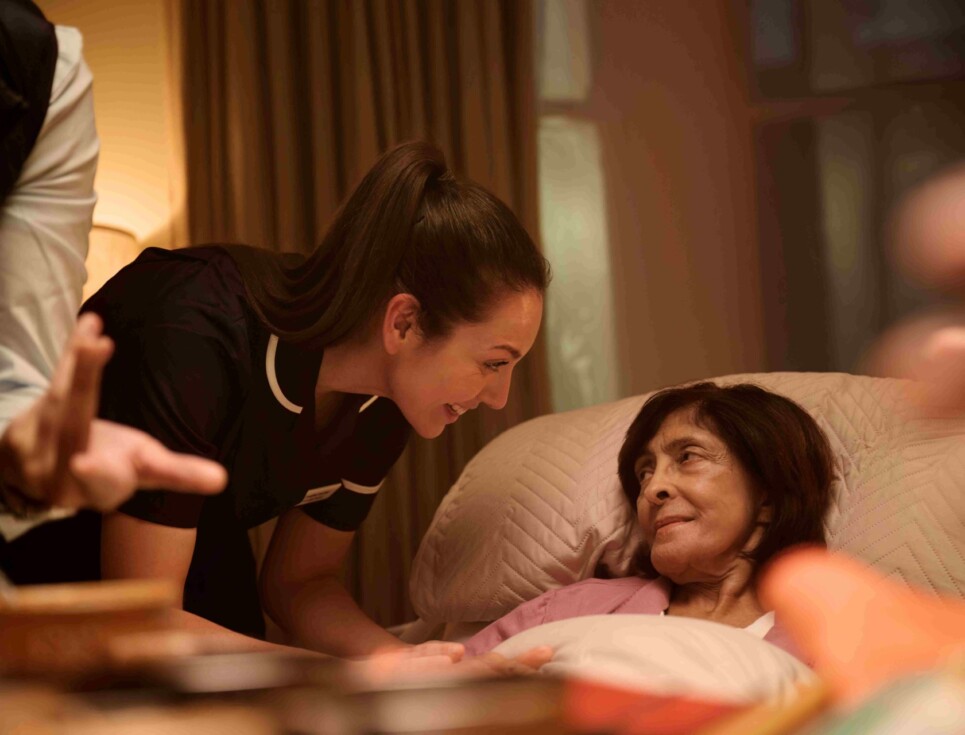 A nurse leans over to speak to a patient lying in bed.